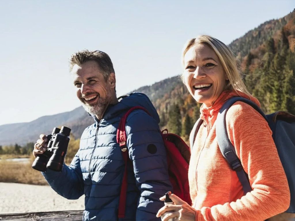Ein lächelnder Mann und eine lächelnde Frau mit Rucksäcken im Freien; der Mann hält ein Fernglas in der Hand, im Hintergrund sind Bäume und Berge zu sehen.