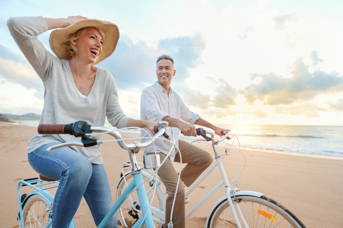 Ein lächelndes Paar fährt bei Sonnenuntergang mit dem Fahrrad an einem Sandstrand entlang und genießt gemeinsam einen entspannten und fröhlichen Moment.