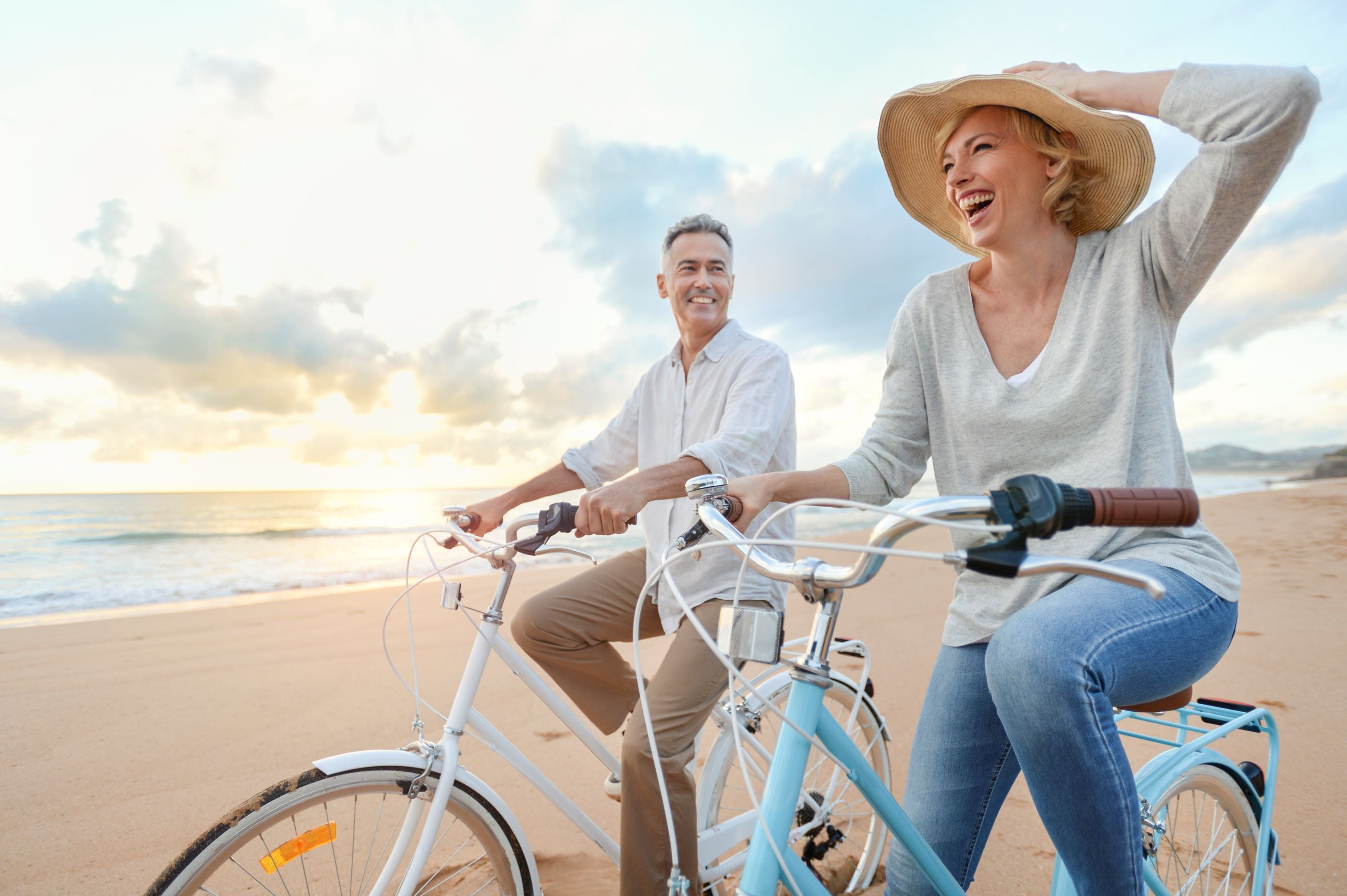 Ein lächelndes Paar fährt bei Sonnenuntergang mit dem Fahrrad an einem Sandstrand entlang; die Frau hält ihren Hut fest, beide wirken entspannt und glücklich.