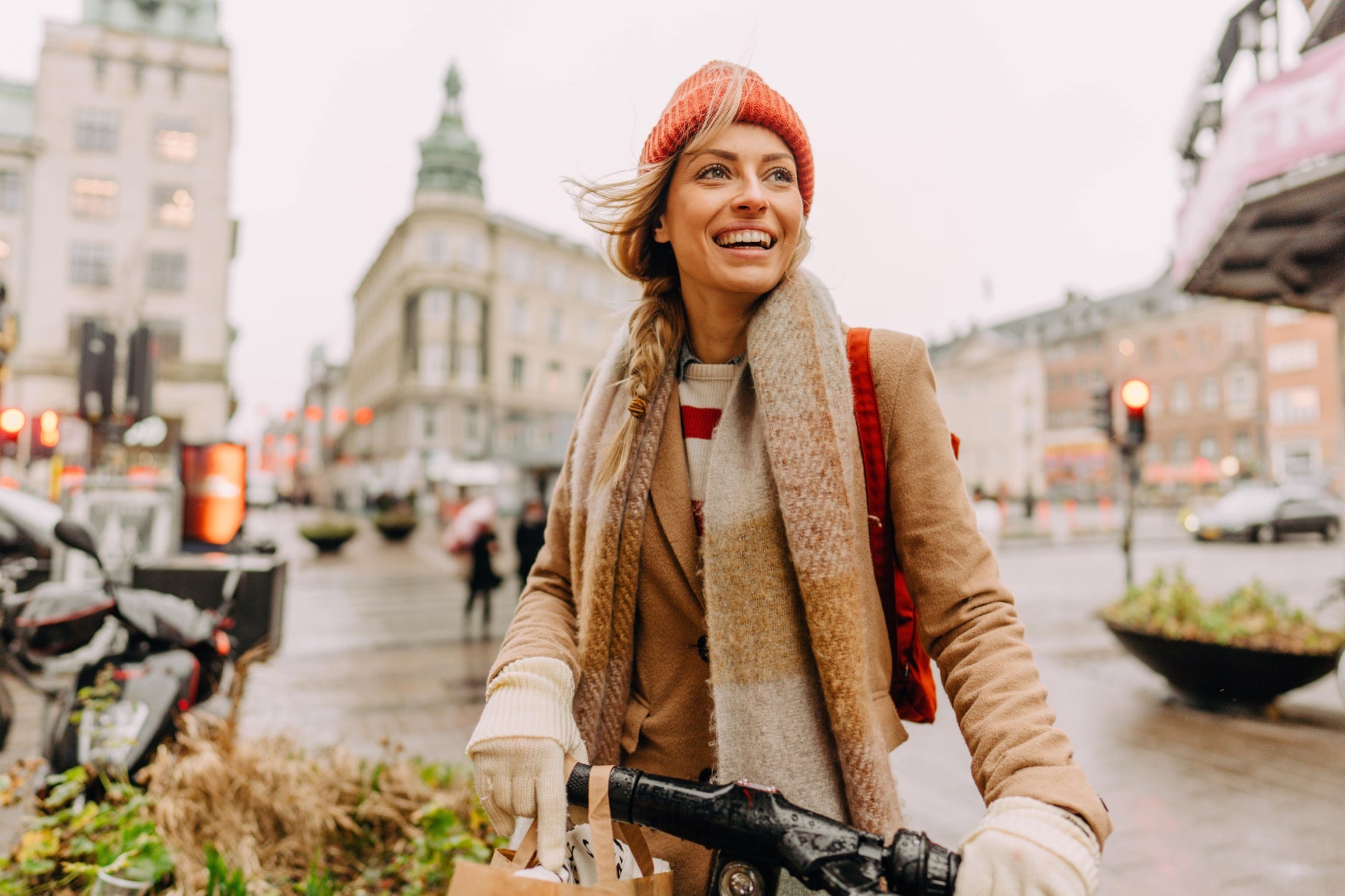 Eine lächelnde Frau mit rotem Hut und Schal fährt an einem kalten Tag mit dem Fahrrad durch eine Stadtstraße.
