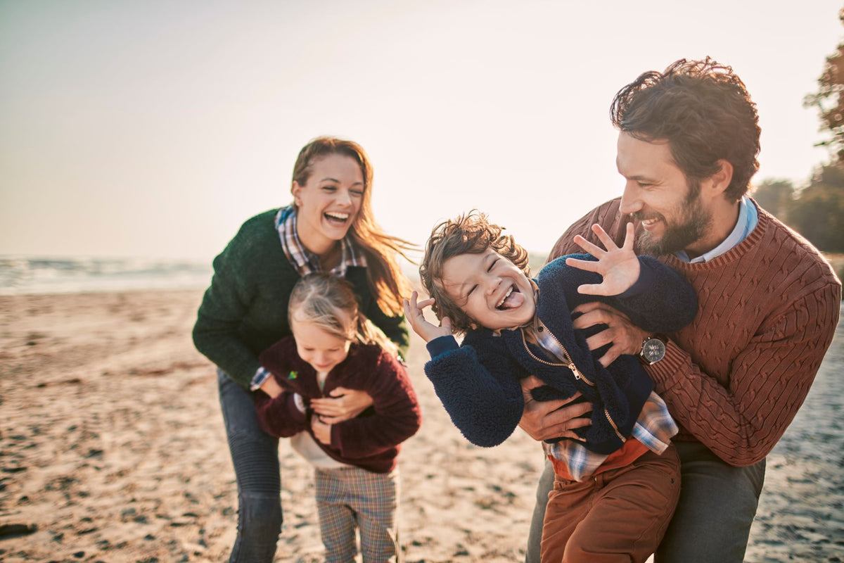 Eine vierköpfige Familie lacht und spielt gemeinsam an einem sonnigen Strand.