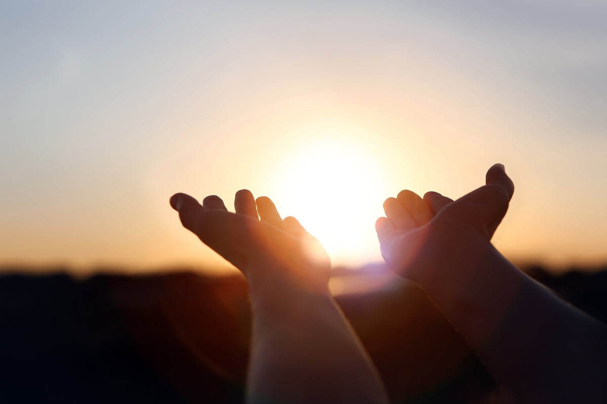 Vor einem verschwommenen Außenhintergrund werden zwei Hände in Richtung einer strahlenden untergehenden Sonne erhoben.