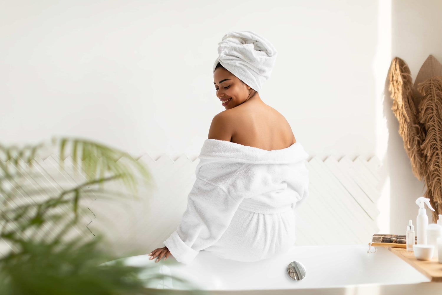 Eine Frau in einem weißen Bademantel und mit einem Handtuch sitzt lächelnd auf einer Badewanne in einem hellen, entspannenden Badezimmer.
