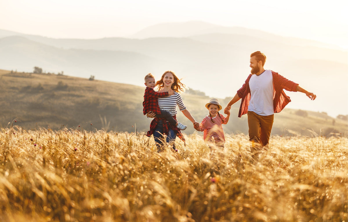 Eine vierköpfige Familie rennt fröhlich durch ein goldenes Feld, im Hintergrund sind Hügel zu sehen, während die Sonne untergeht.