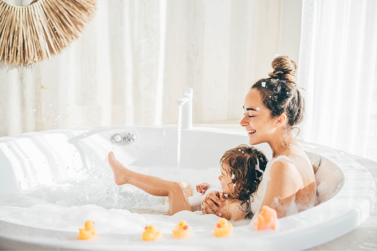 Eine lächelnde Frau und ein Kind genießen gemeinsam ein Schaumbad in einem hellen Badezimmer, in dem Gummienten am Rand der Badewanne liegen.