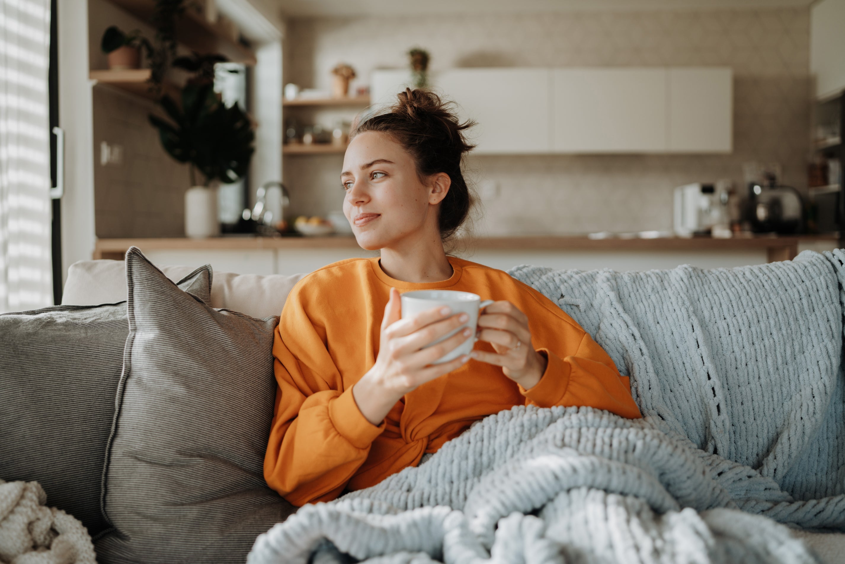 Eine Frau in einem orangefarbenen Pullover sitzt auf einem Sofa, hält eine Tasse in der Hand und blickt nachdenklich aus dem Fenster.