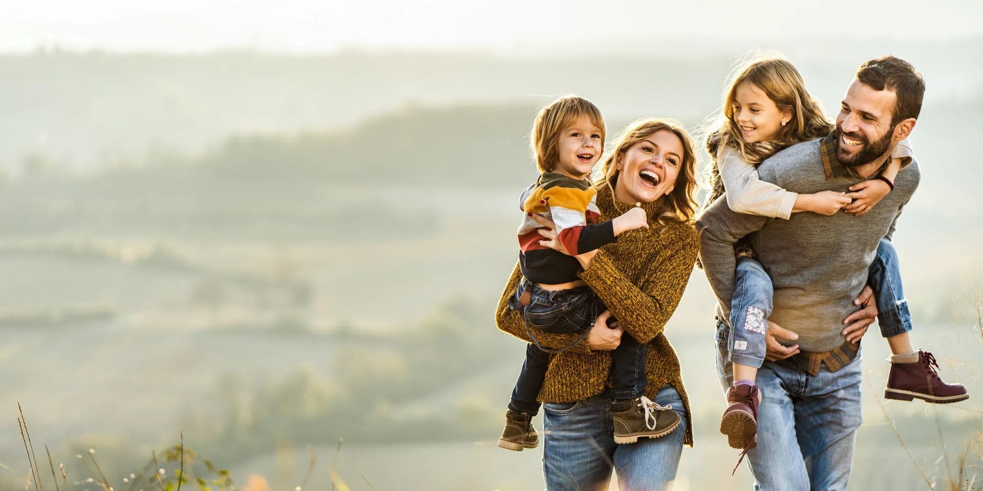 Eine lächelnde vierköpfige Familie genießt die Zeit im Freien; die Eltern tragen ihre Kinder auf dem Rücken auf einer sonnigen, weiten Wiese.