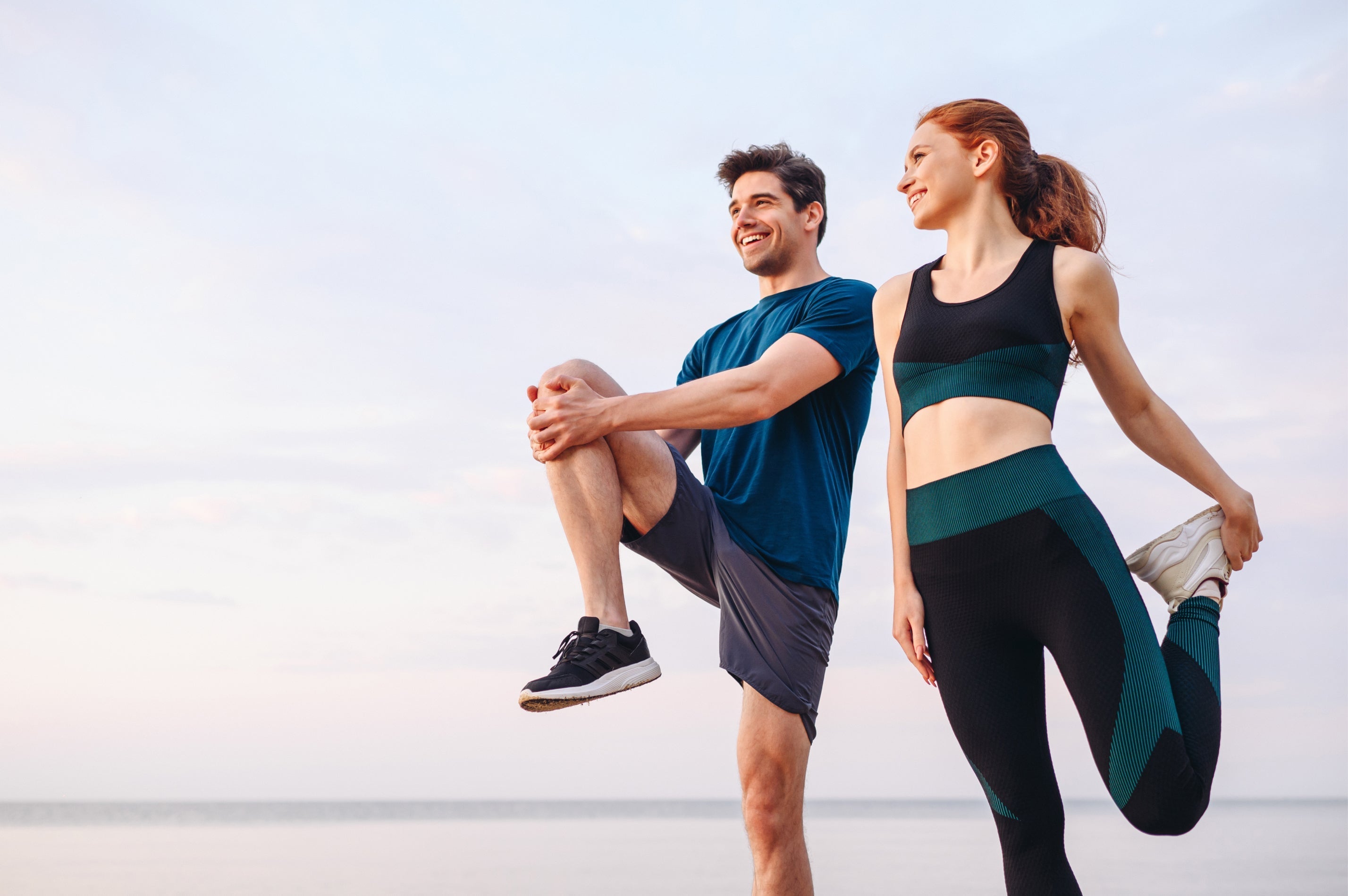 Zwei Personen in Sportkleidung, die lächelnd gemeinsam draußen am Wasser Dehnübungen machen.