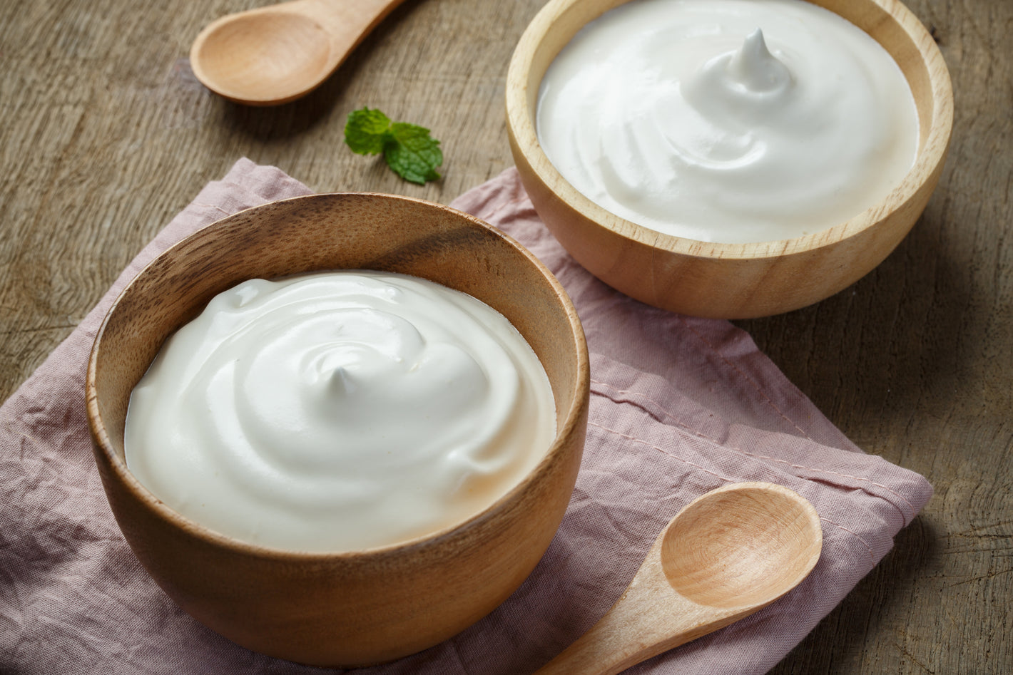 Zwei Holzschalen mit cremigem Joghurt auf einem rosa Tischtuch, dazu Minzblätter und Holzlöffel auf einem Holztisch.
