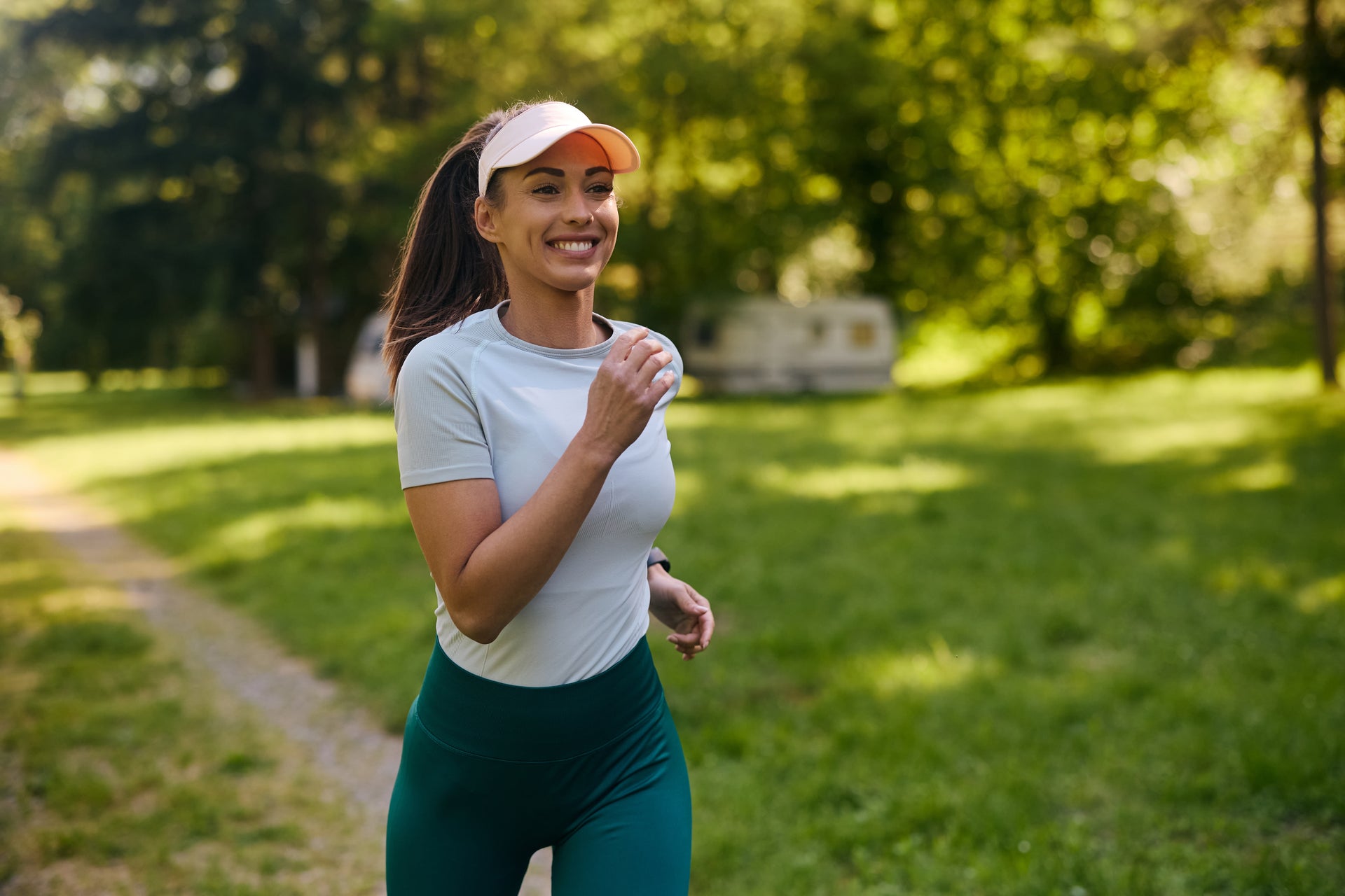Eine lächelnde Frau, die auf einem sonnigen Weg in einem grünen Park joggt; sie trägt eine Schirmmütze und Sportkleidung.