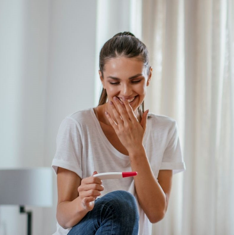 Eine lächelnde Frau sitzt in einem Raum, hält einen Schwangerschaftstest in der Hand und hält sich vor Überraschung und Freude die Hand vor den Mund.