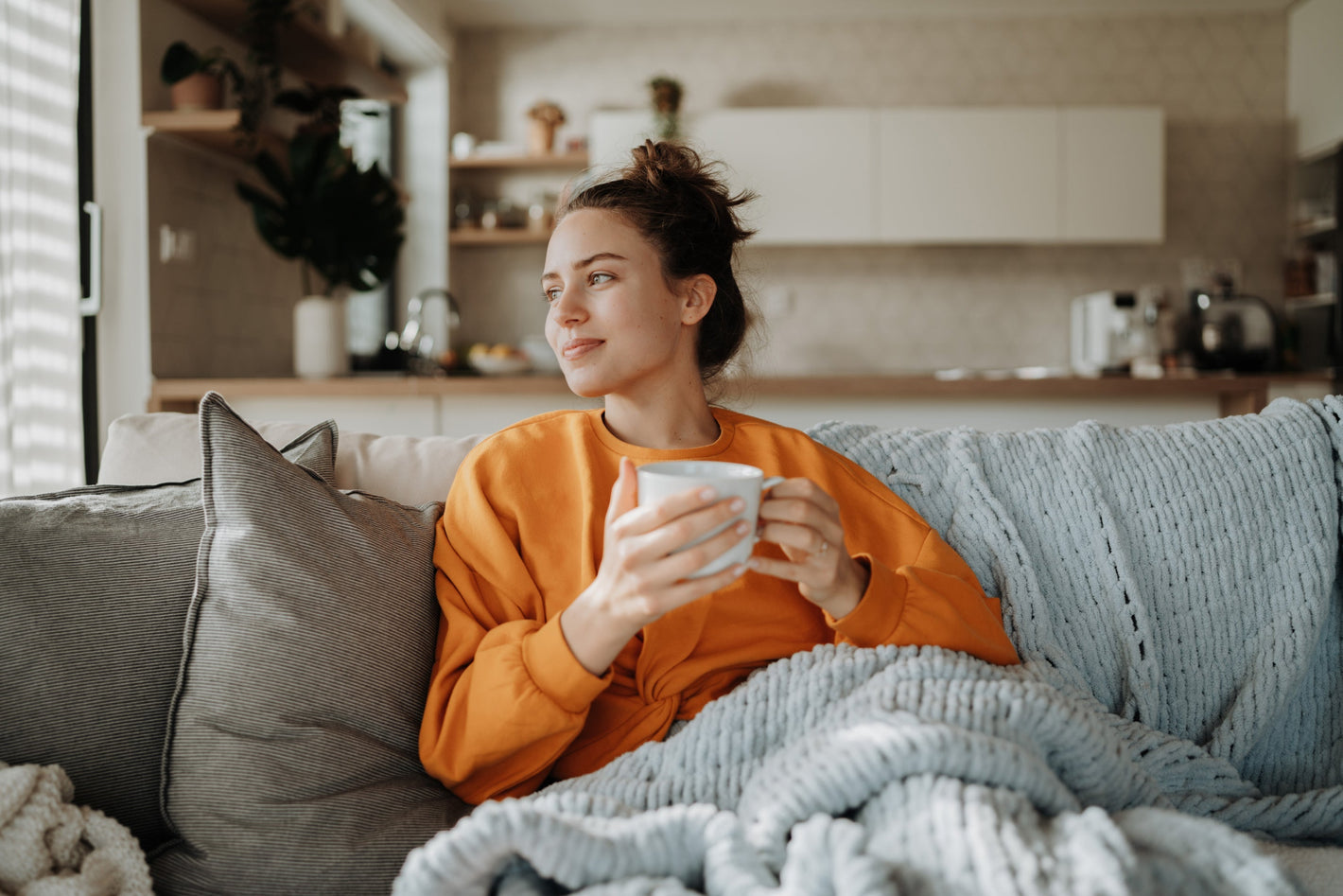 Eine Frau in einem orangefarbenen Sweatshirt sitzt mit einer Tasse auf einem Sofa, in eine Decke gehüllt, und schaut aus dem Bild heraus.